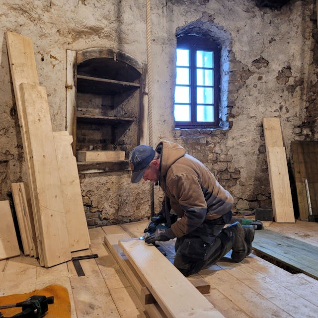 Ein Arbeiter kniet auf dem Boden, er schneidet ein Fußbodenbrett zurecht. An den Wänden lehnen unterschiedlich große Bretter.