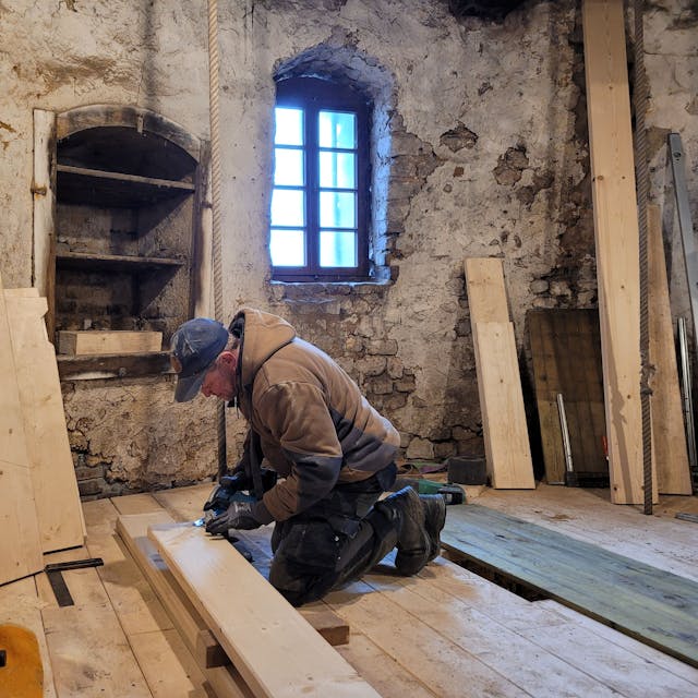 Ein Arbeiter kniet auf dem Boden, er schneidet ein Fußbodenbrett zurecht. An den Wänden lehnen unterschiedlich große Bretter.
