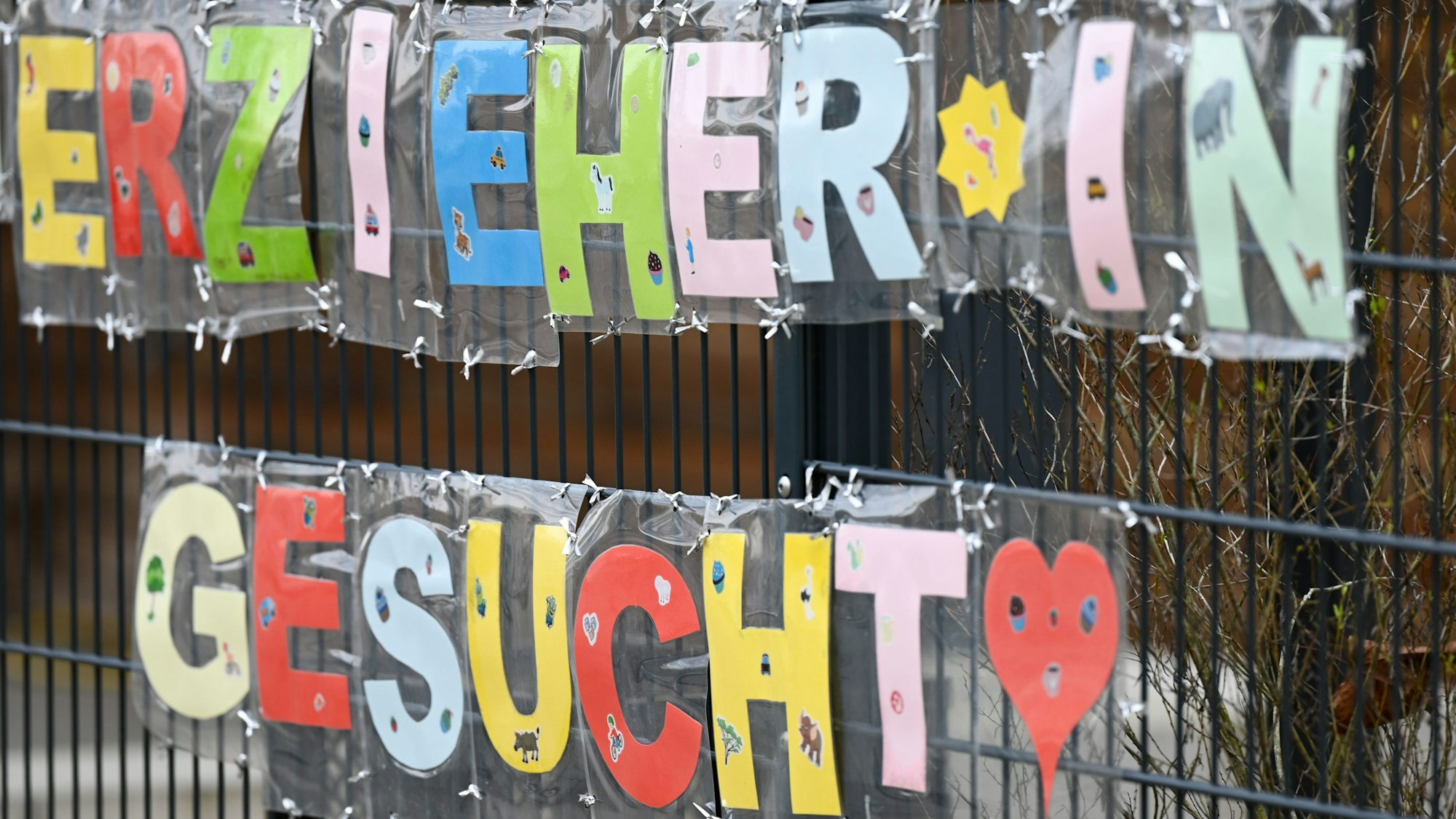 Mit bunten Buchstaben sind die Worte „Erzieher*in gesucht“ geformt, die an einem Zaun einer Kindertagesstätte. (Symbolbild)