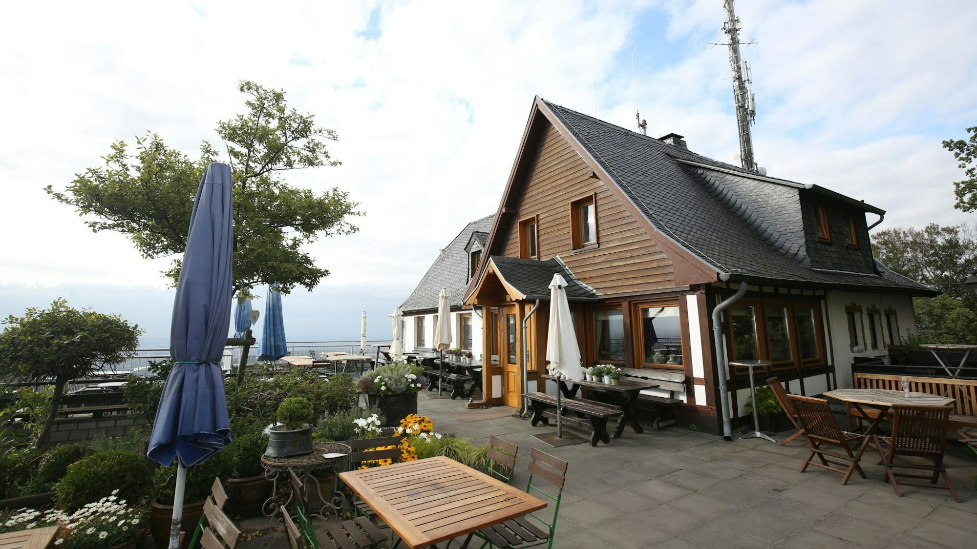 Ein holzverkleidetes Gasthaus, vor dem Holztische und Sonnenschirme stehen. Die Silhouette des Siebengebirges ist zu erkennen.