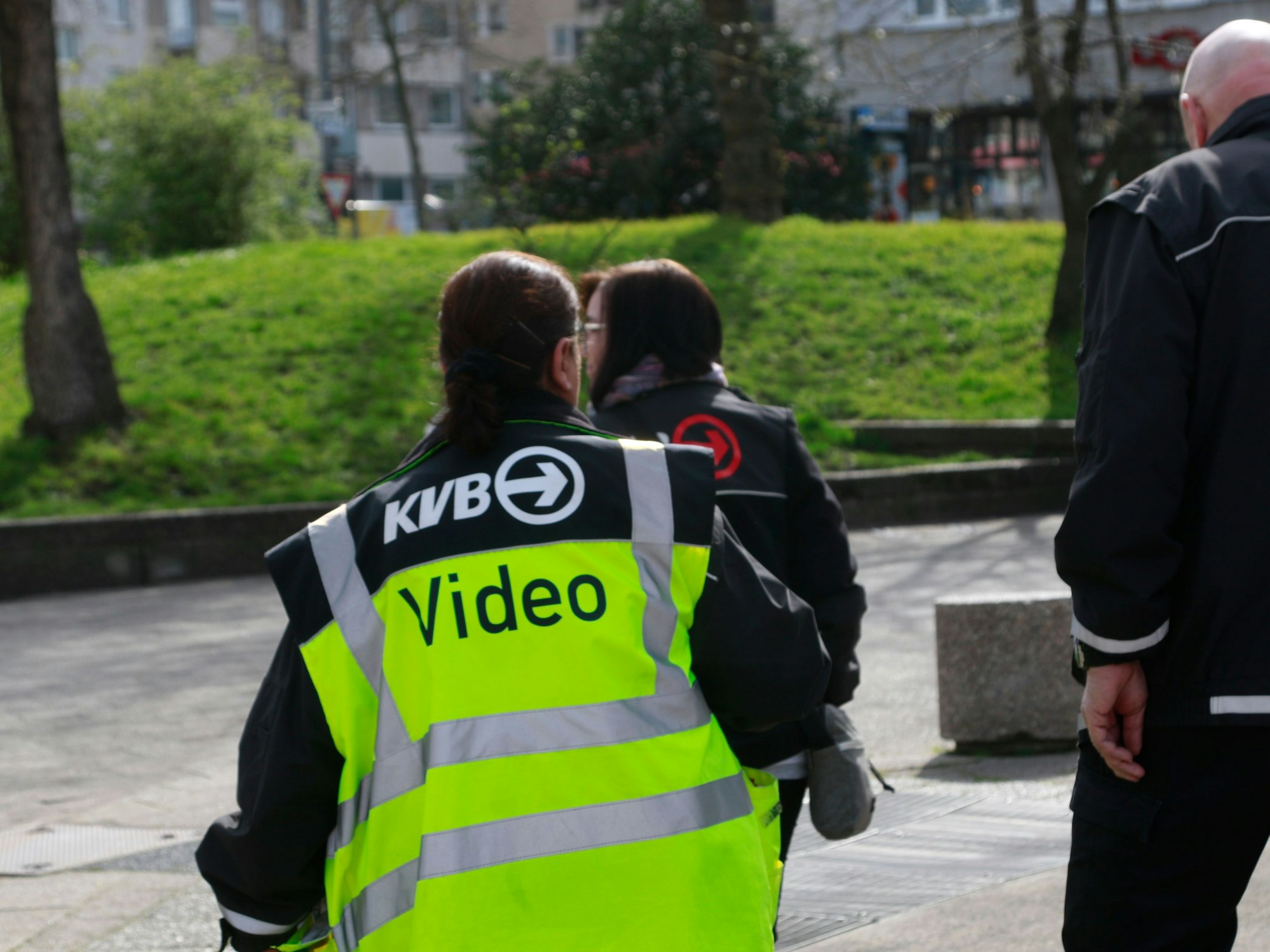 Drei Personen gehen über einen Platz, eine Frau trägt eine Weste mit der Aufschrift „KVB Video“.