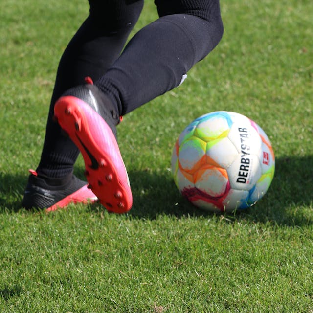 Die Füße eines Fußballspielers in pink-schwarzen Schuhen sind zu sehen, kurz bevor er einen Ball schießt.
