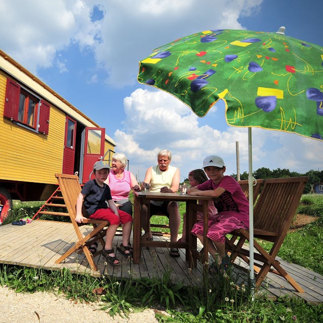 Ein gelber Zirkuswagen, davor sitzen Großeltern mit drei Kindern unter einem Sonnenschirm am Tisch.