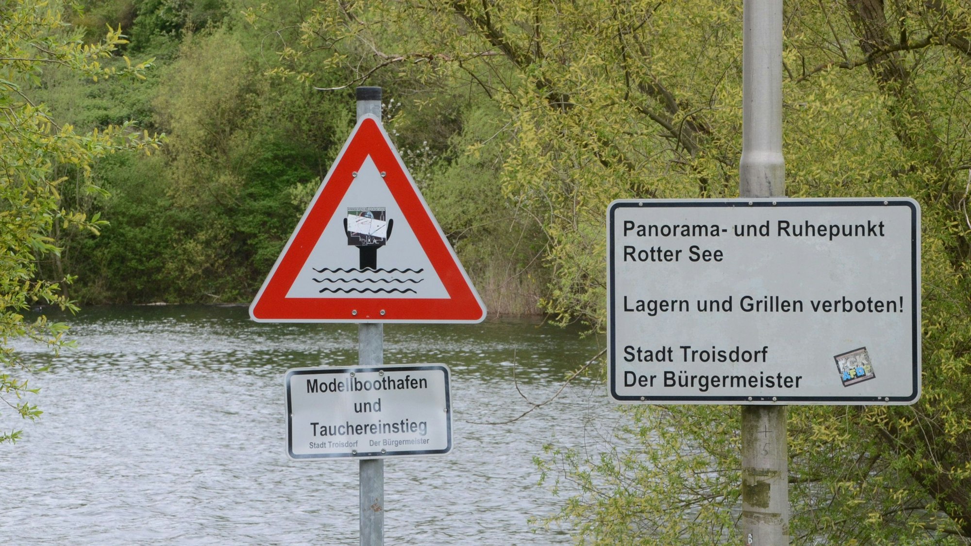 Schilder an einem Seeufer. „Panorama- und Ruhepunkt Rotter See“ ist zu lesen, außerdem „Lagern und Grillen verboten“. Ein anderes Schild weist das Gelände als Tauchereinstieg und Modellboothafen aus.