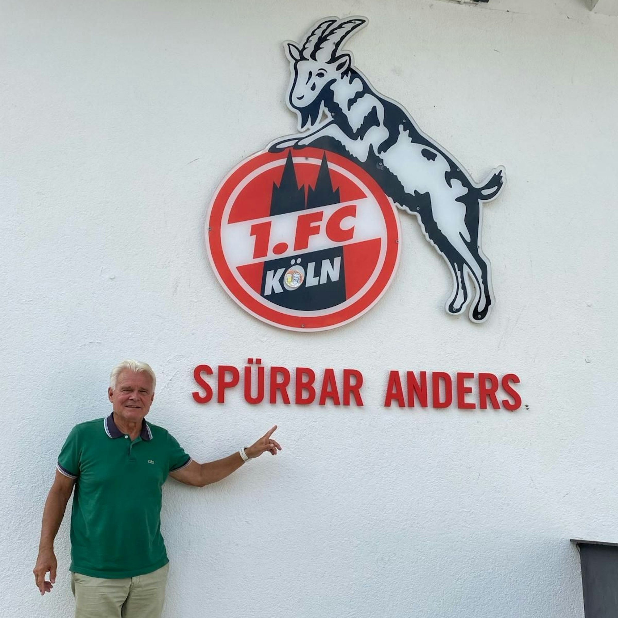 Rolf Birkhölzer zeigt auf eine Wand, auf der das Logo des 1. FC Köln angebracht ist.