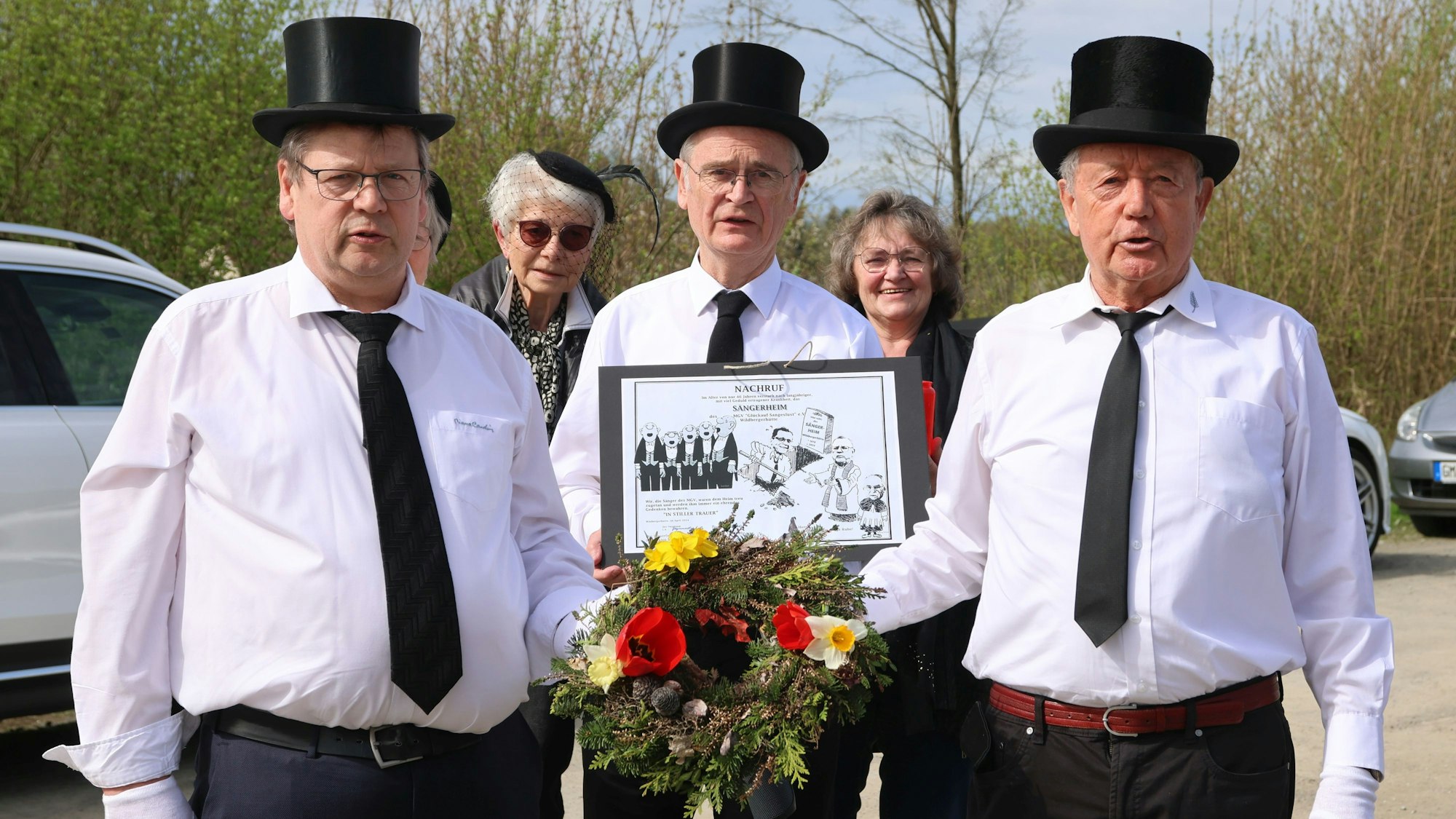 Drei Herren mit schwarzen Zylindern tragen ein Blumengesteck.