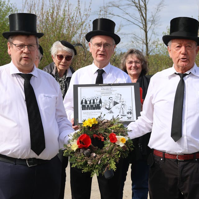 Drei Herren mit schwarzen Zylindern tragen ein Blumengesteck.