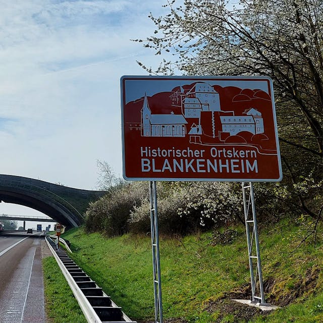Am rechten Fahrbahnrand steht das neue Schild, auf dem zu lesen ist: „Historischer Ortskern Blankenheim“. Im Hintergrund ist die Wildbrücke zu sehen.