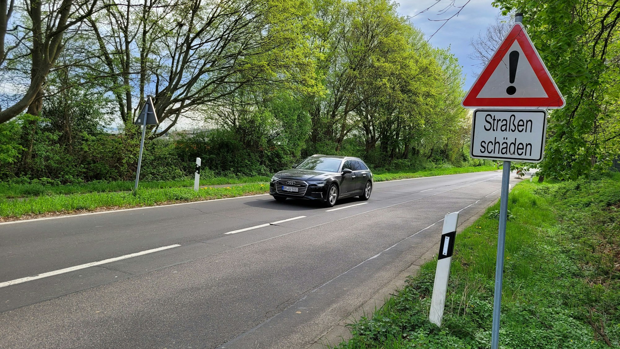 Eine zweispurige Straße, ein Auto auf dem linken Fahrstreifen, rechts ein Hinweisschild mit dem Schriftzug „Straßenschäden“.