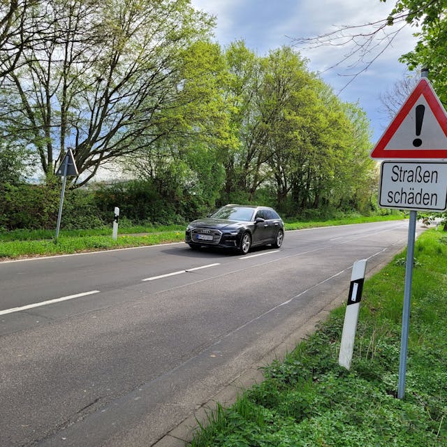 Eine zweispurige Straße, ein Auto auf dem linken Fahrstreifen, rechts ein Hinweisschild mit dem Schriftzug „Straßenschäden“.