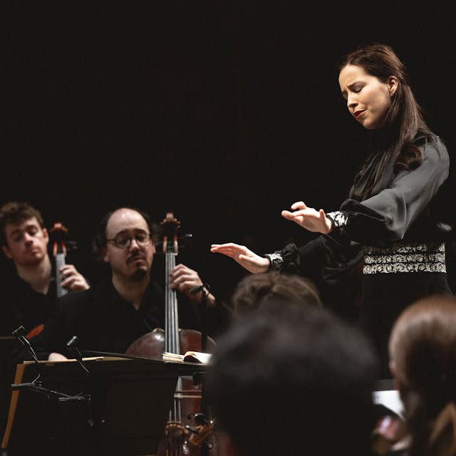Auf dem Bild ist eine junge Dirigentin mit Musikern zu sehen.