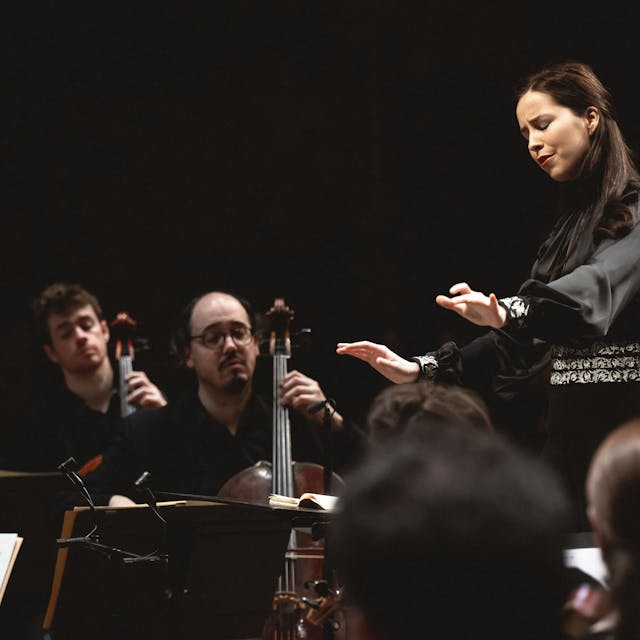 Auf dem Bild ist eine junge Dirigentin mit Musikern zu sehen.