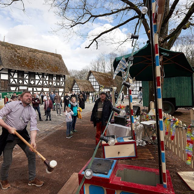 Ein Besucher des historischen Jahrmarkts schwingt den Hammer beim „Hau den Lukas“. Im Hintergrund stehen Fachwerkhäuser.