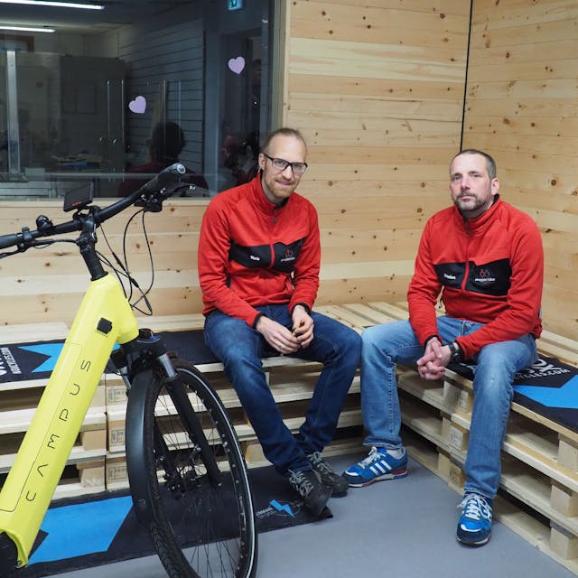 Zwei Männer sitzen auf Bänken, die aus Holz-Paletten gebaut sind. Vor ihnen steht ein gelbes Fahrrad.