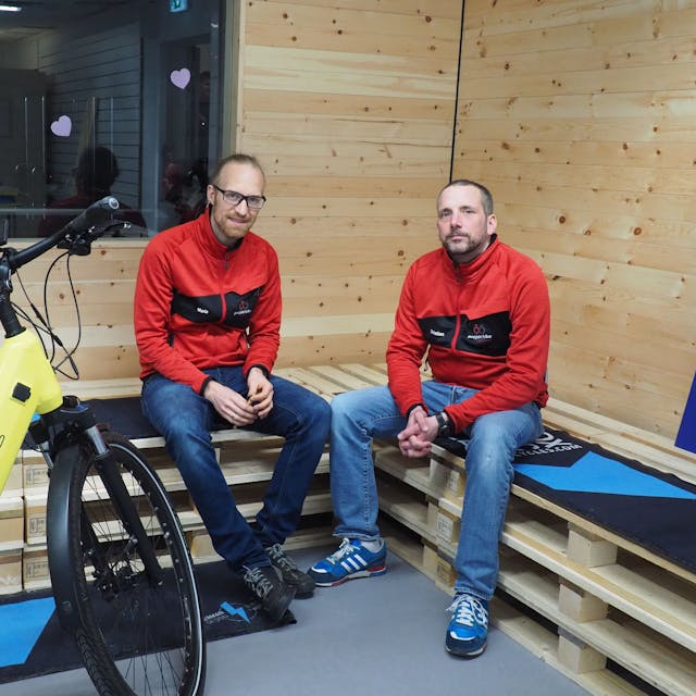 Zwei Männer sitzen auf Bänken, die aus Holz-Paletten gebaut sind. Vor ihnen steht ein gelbes Fahrrad.