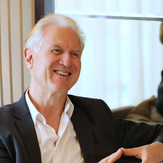 Chefdirigent Christoph Poppen sitzt auf einem Sessel und blickt freundlich lächelnd. Er trägt ein weißes Hemd und ein schwarzes Sakko.