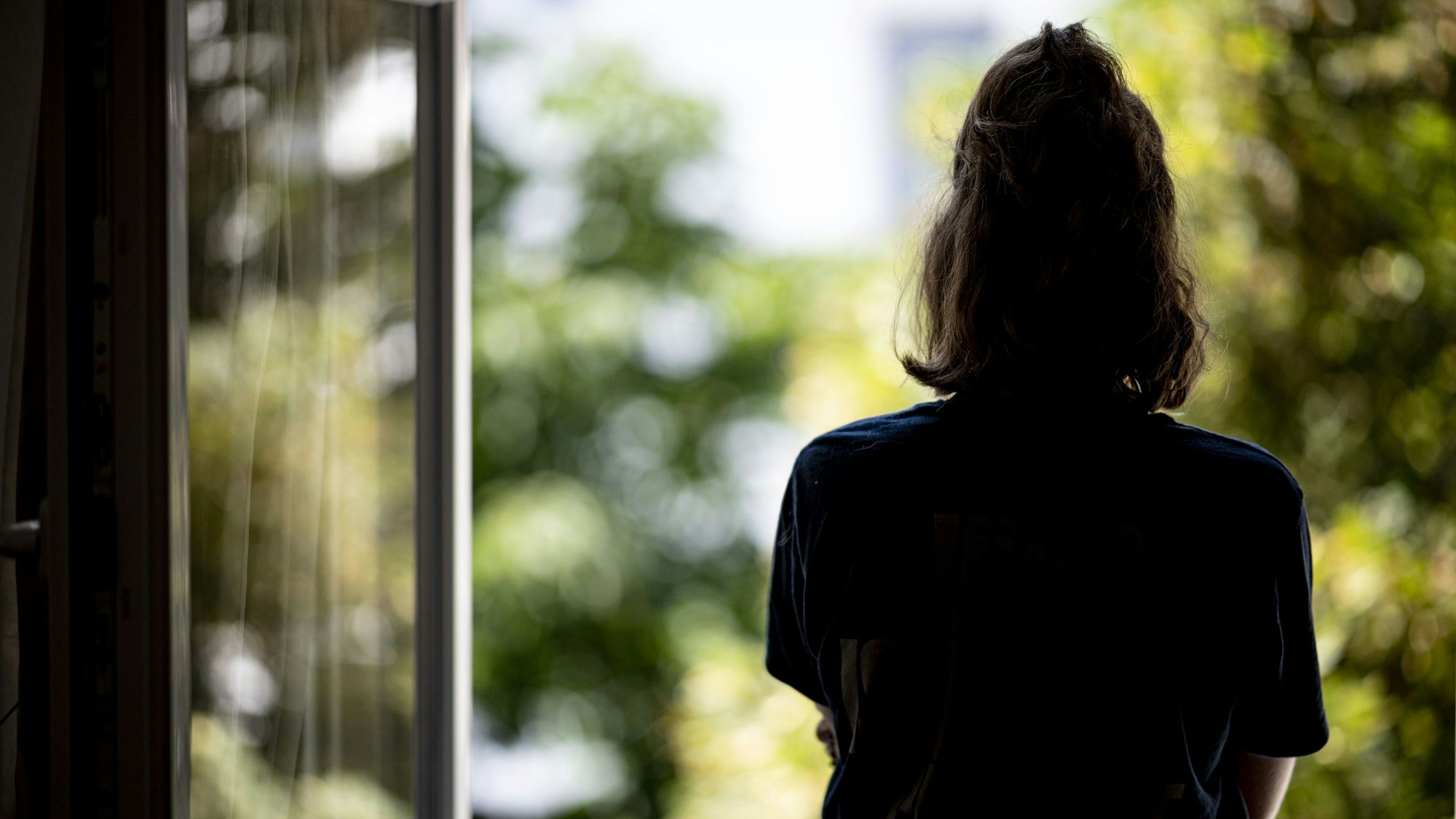Eine Frau steht allein in ihrer Wohnung an einem Fenster und schaut ins Freie.