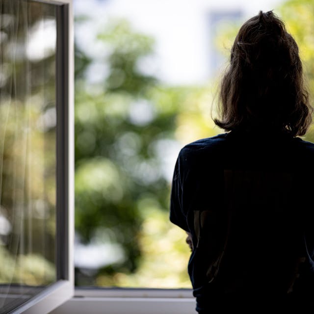 Eine Frau steht allein in ihrer Wohnung an einem Fenster und schaut ins Freie.