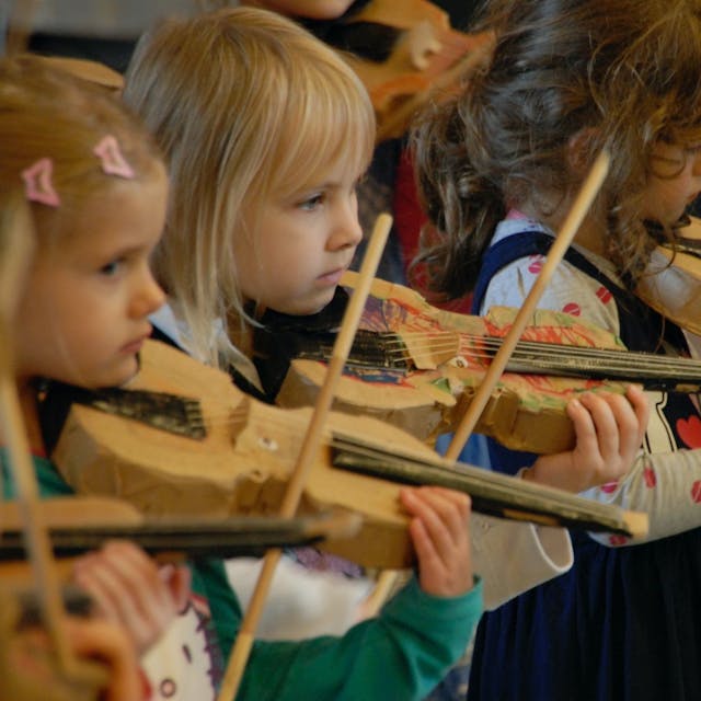 Drei Mädchen spielen Geige mit einem Pappinstrument.