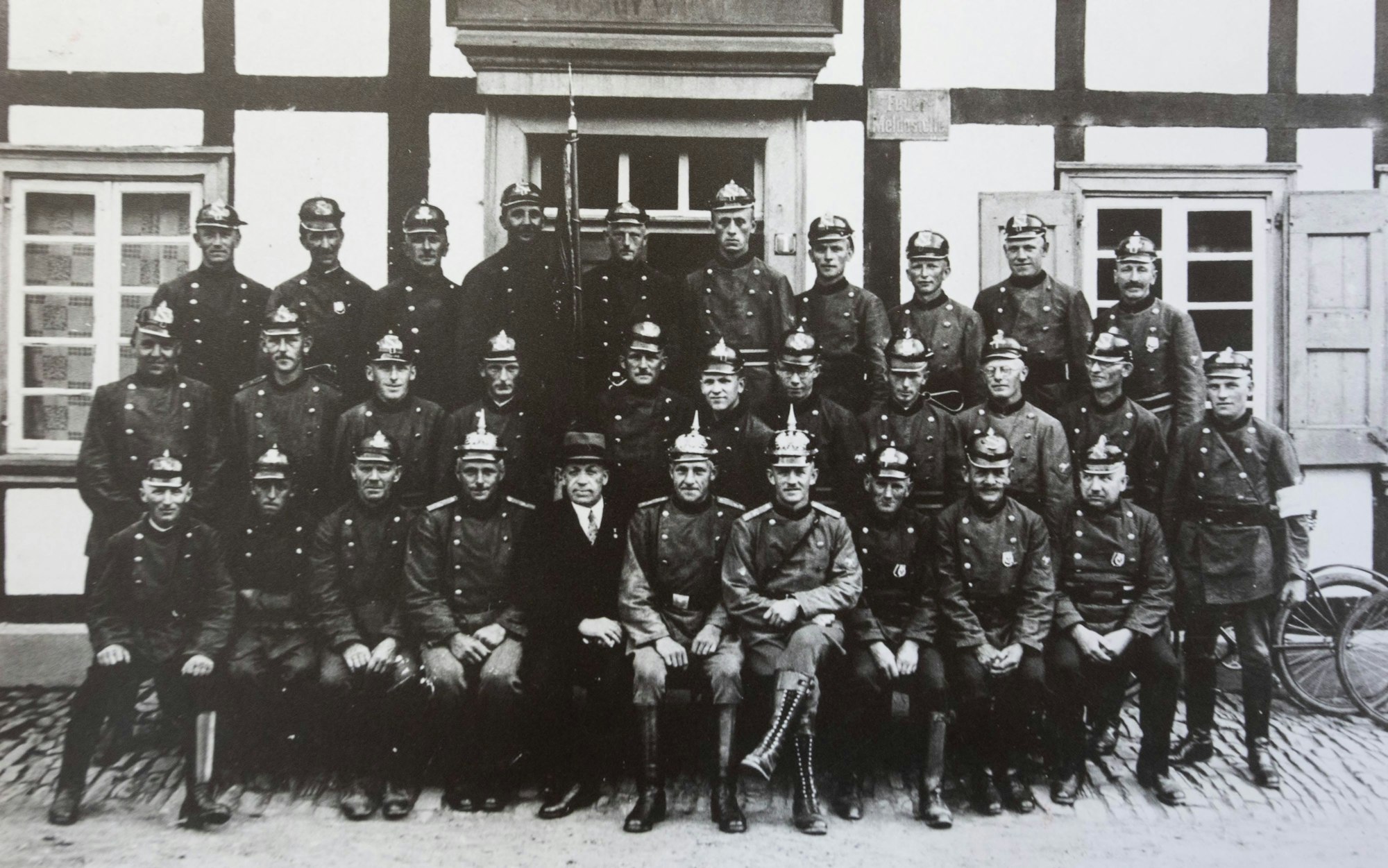 Schwarz-weiß-Gruppenbild von Männern vor einem Fachwerkhaus. Sie tragen Uniformen und preußische Pickelhauben.