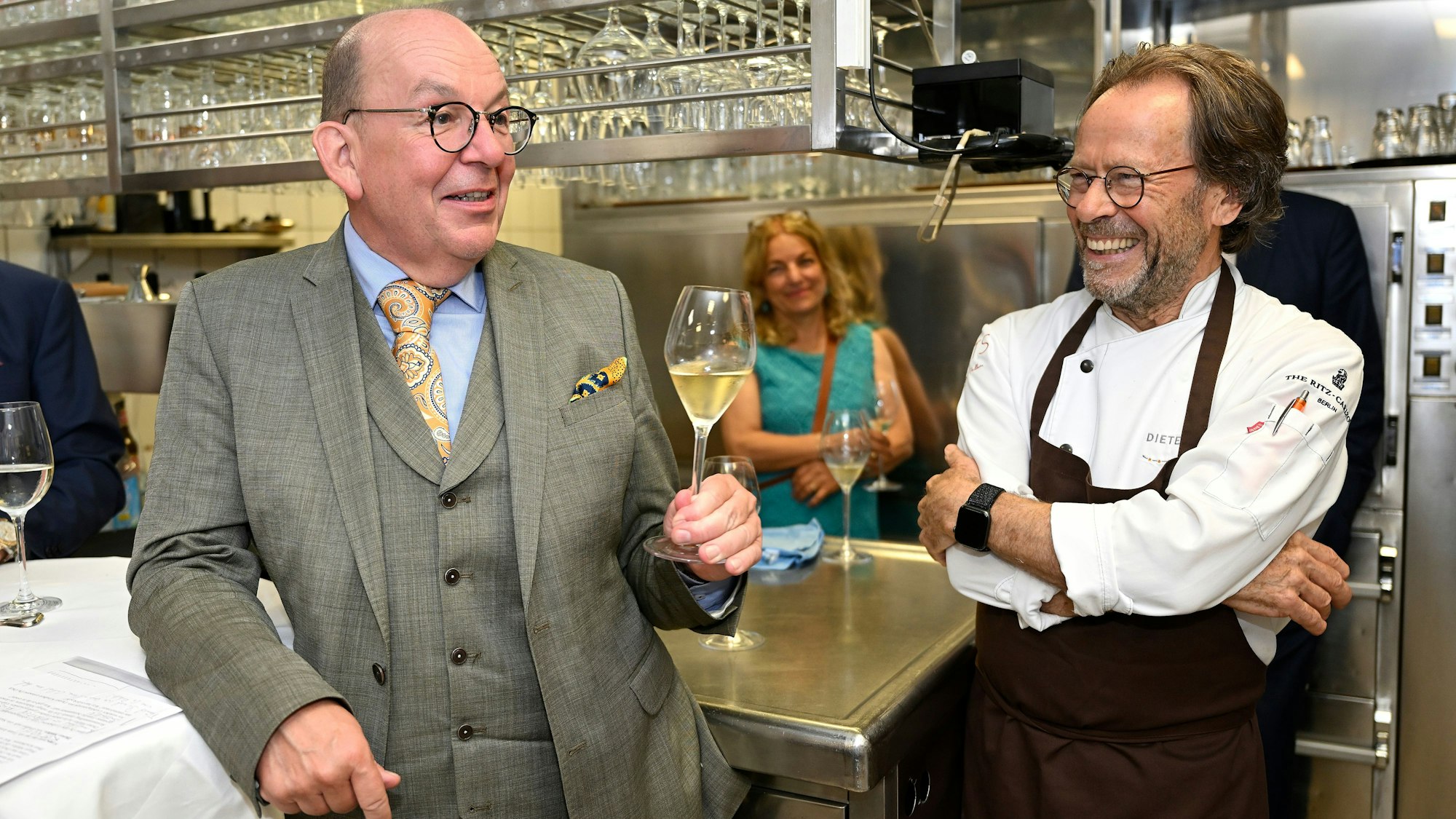 Ein Koch unterhält sich lachend mit einem Gast in der Küche, der ein Weinglas in der Hand hält.