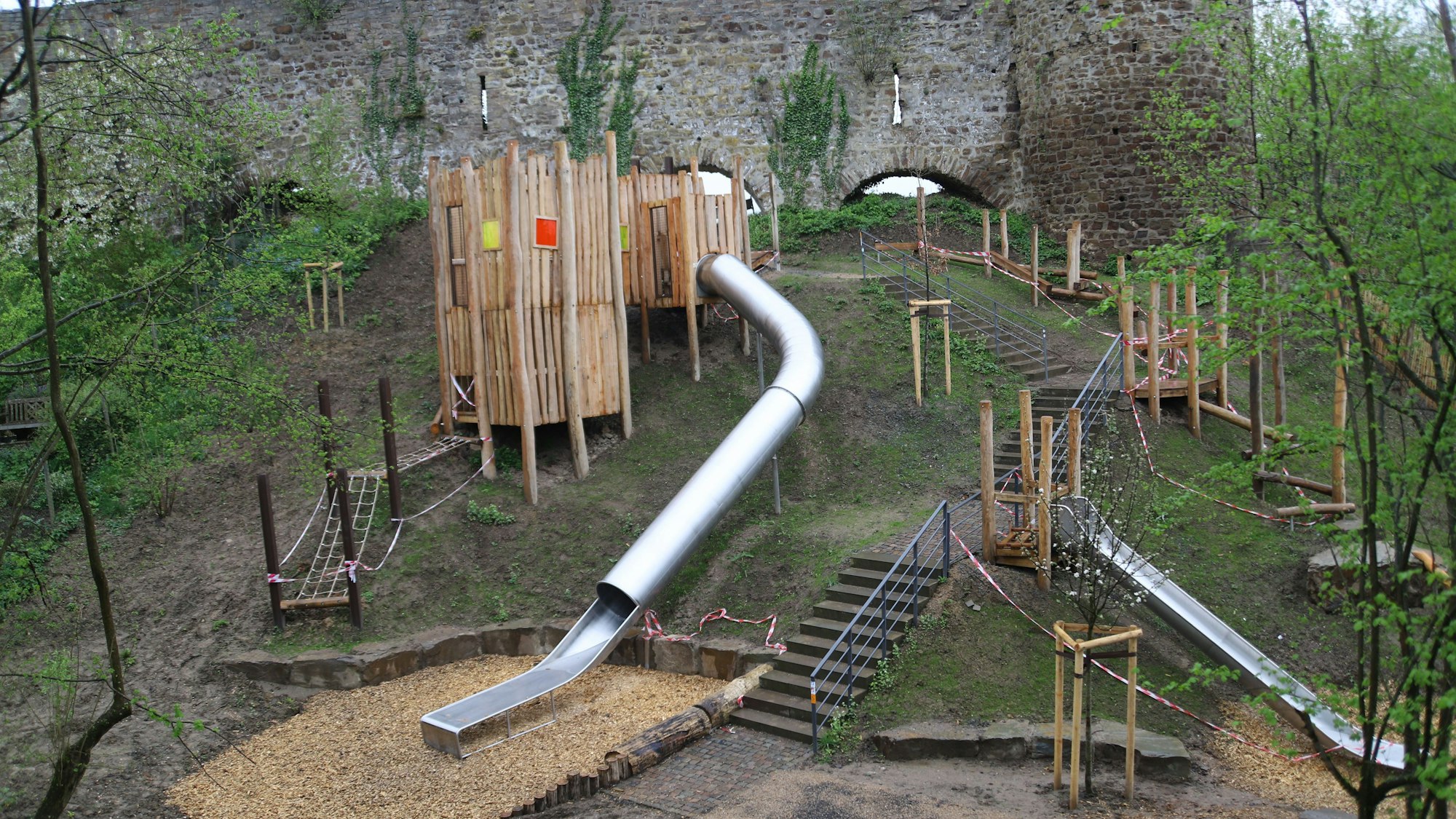 Das Bild zeigt einen Spielplatz an einem Steilhang mit zwei Rutschbahnen, einem Holzturm, einer Treppe und Sitzsteinen.