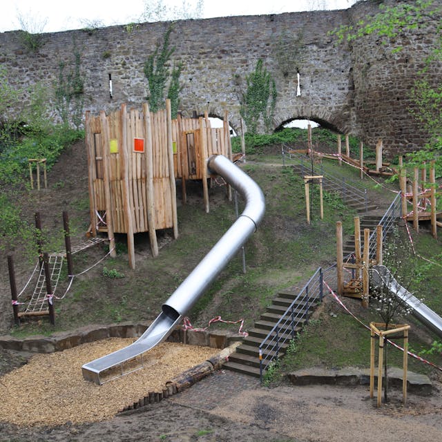 Das Bild zeigt einen Spielplatz an einem Steilhang mit zwei Rutschbahnen, einem Holzturm, einer Treppe und Sitzsteinen.