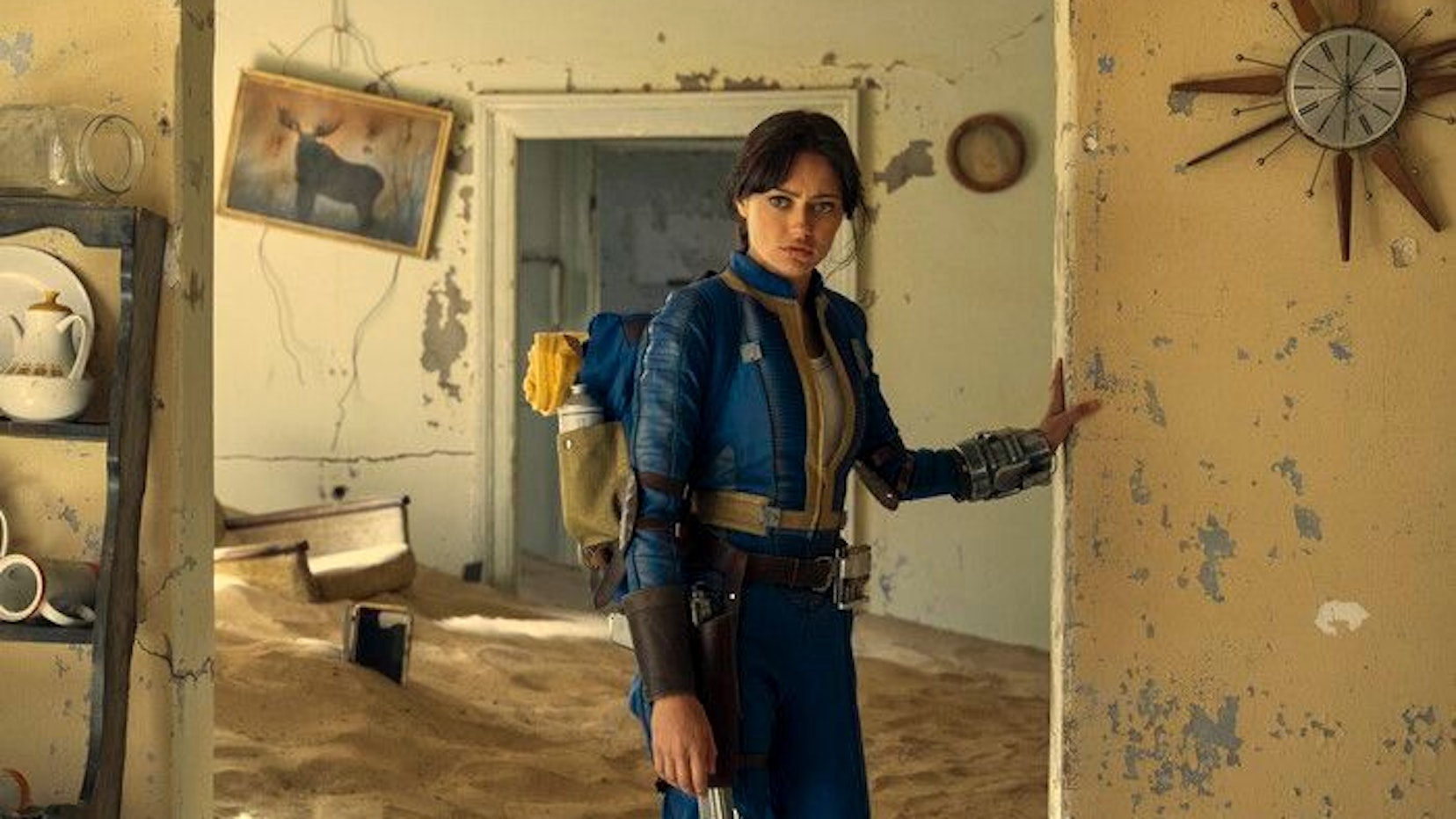 Lucy trägt einen blauen Overall mit gelben Akzenten. Sie lehnt sich im zerstörten inneren eines verwitterten Hauses mit der Hand an eine Wand.