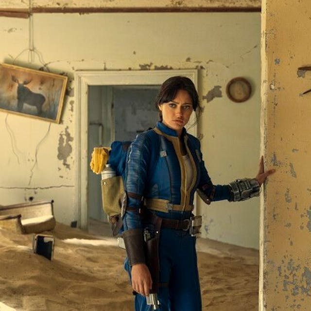 Lucy trägt einen blauen Overall mit gelben Akzenten. Sie lehnt sich im zerstörten inneren eines verwitterten Hauses mit der Hand an eine Wand.&nbsp;