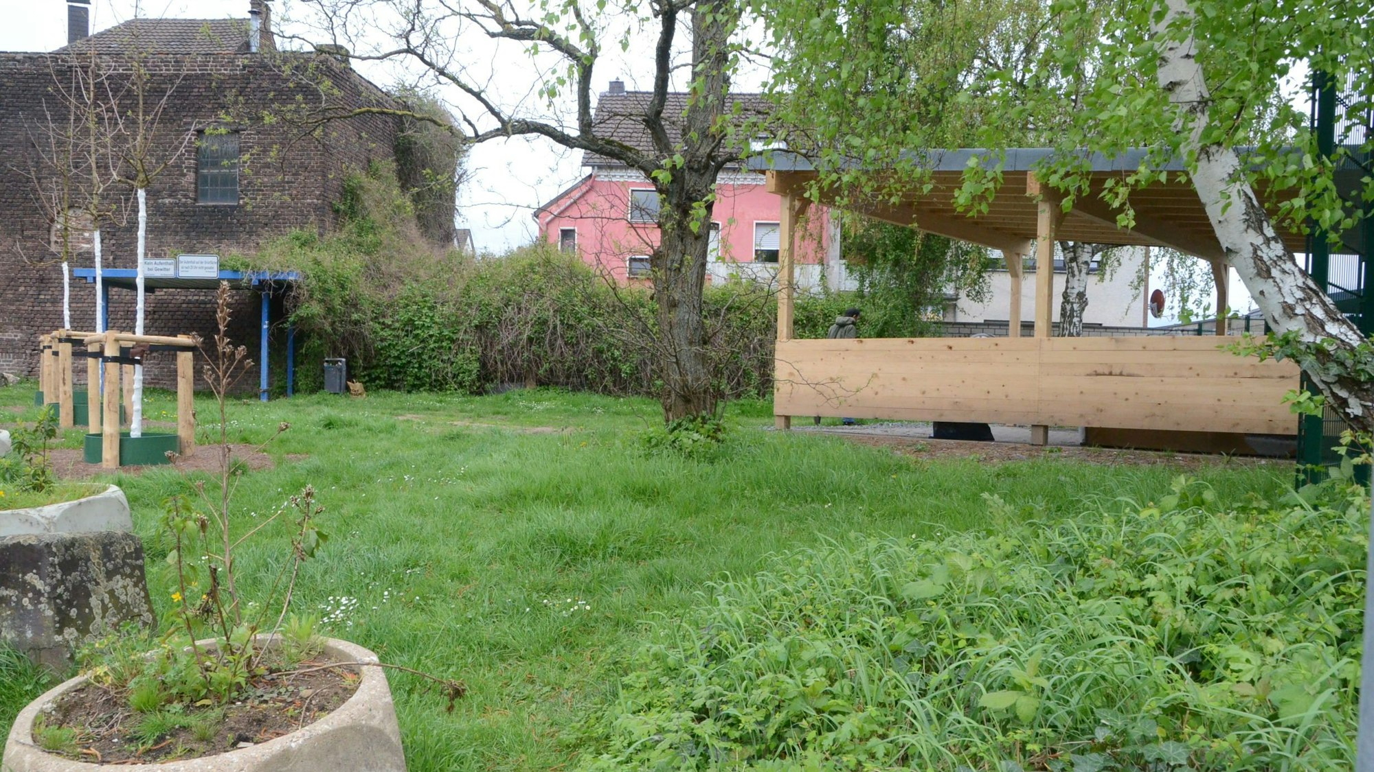 Eine Grünfläche mit Bäumen. Links im Hintergrund steht ein Unterstand aus blauem Metall, rechts eine Art Carport aus Holz mit halbhohen Seitenwänden.