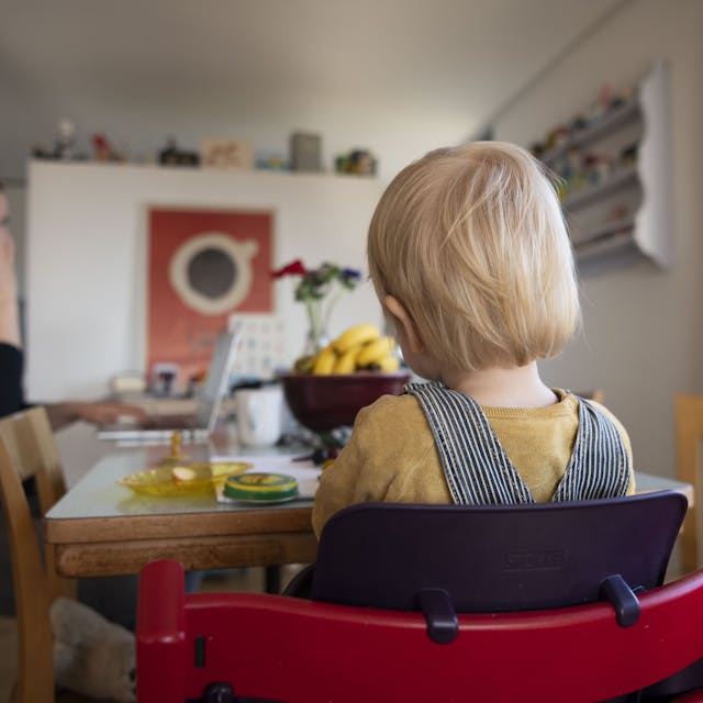 Eine Frau sitzt im Homeoffice an ihrem Laptop und telefoniert, während ihr Kind neben ihr in einem Kinderstuhl am Tisch sitzt.