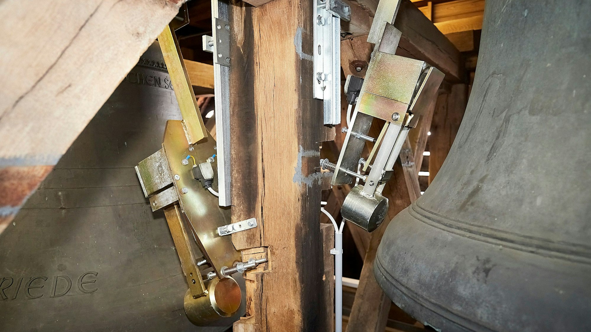 An einem alten Glockenstuhl aus Holz sind neue, pneumatische Schlagwerke angebracht, die eine Glocke, die zum Teil zum rechts im Bild zu sehen ist, zum Klingen bringen.