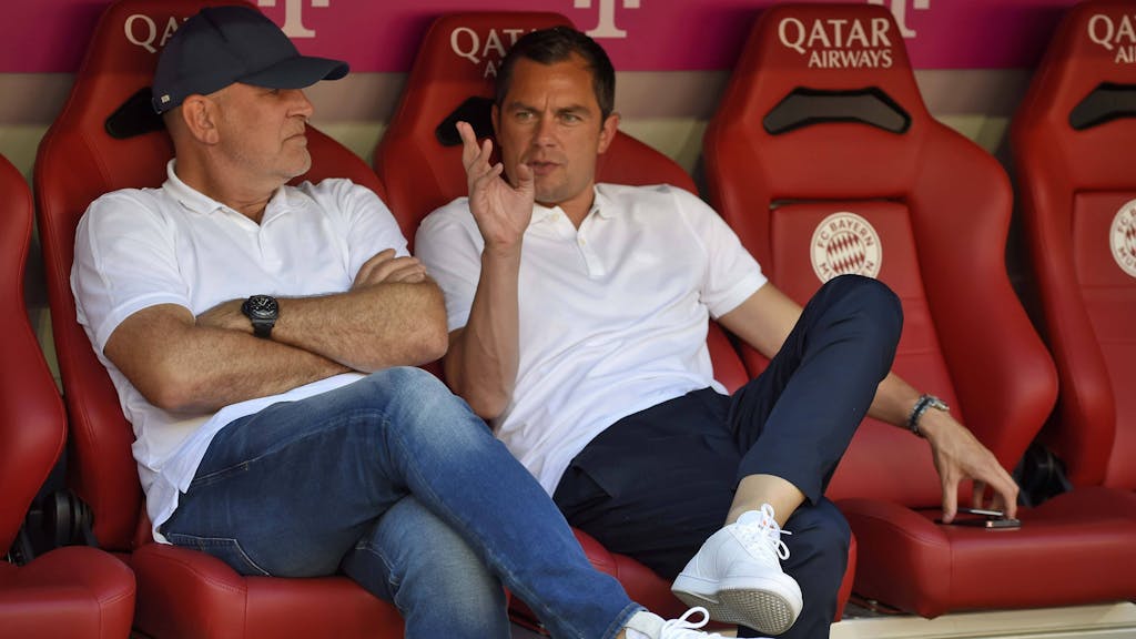 Jörg Schmadtke und Marcel Schäfer sitzen vor dem Spiel beim FC Bayern auf der Bank und sprechen miteinander.