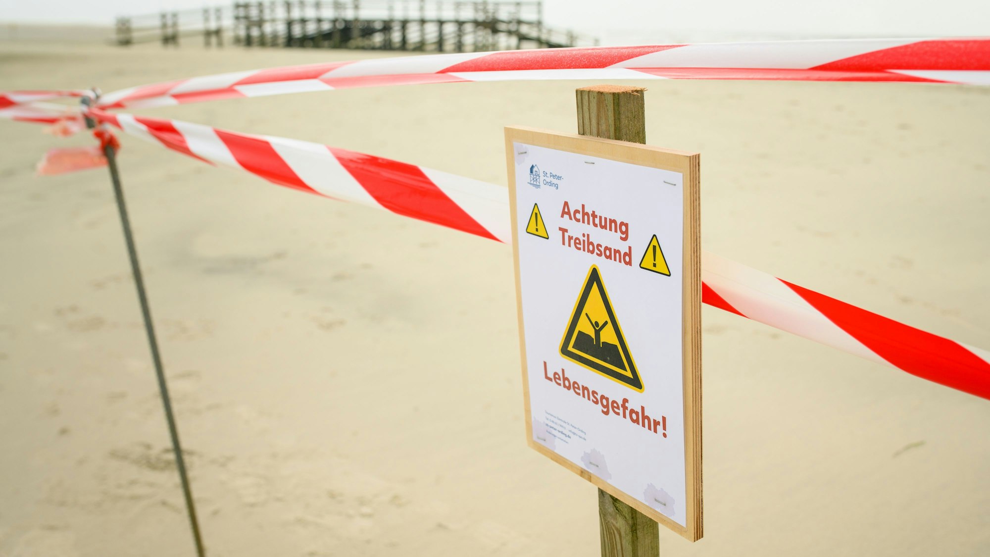Flatterband und ein Warnhinweis („Achtung Treibsand – Lebensgefahr!“) sind an einem abgesperrten Bereich auf dem Strand von St. Peter-Ording zu sehen.