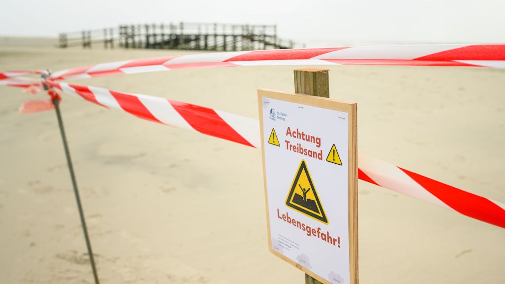 Flatterband und ein Warnhinweis („Achtung Treibsand – Lebensgefahr!“) sind an einem abgesperrten Bereich auf dem Strand von St. Peter-Ording zu sehen.