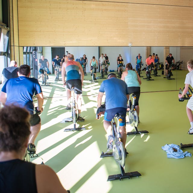 Mehrere Menschen sitzen auf Cycling-Bikes in einem großen Raum vor einem Spiegel.
