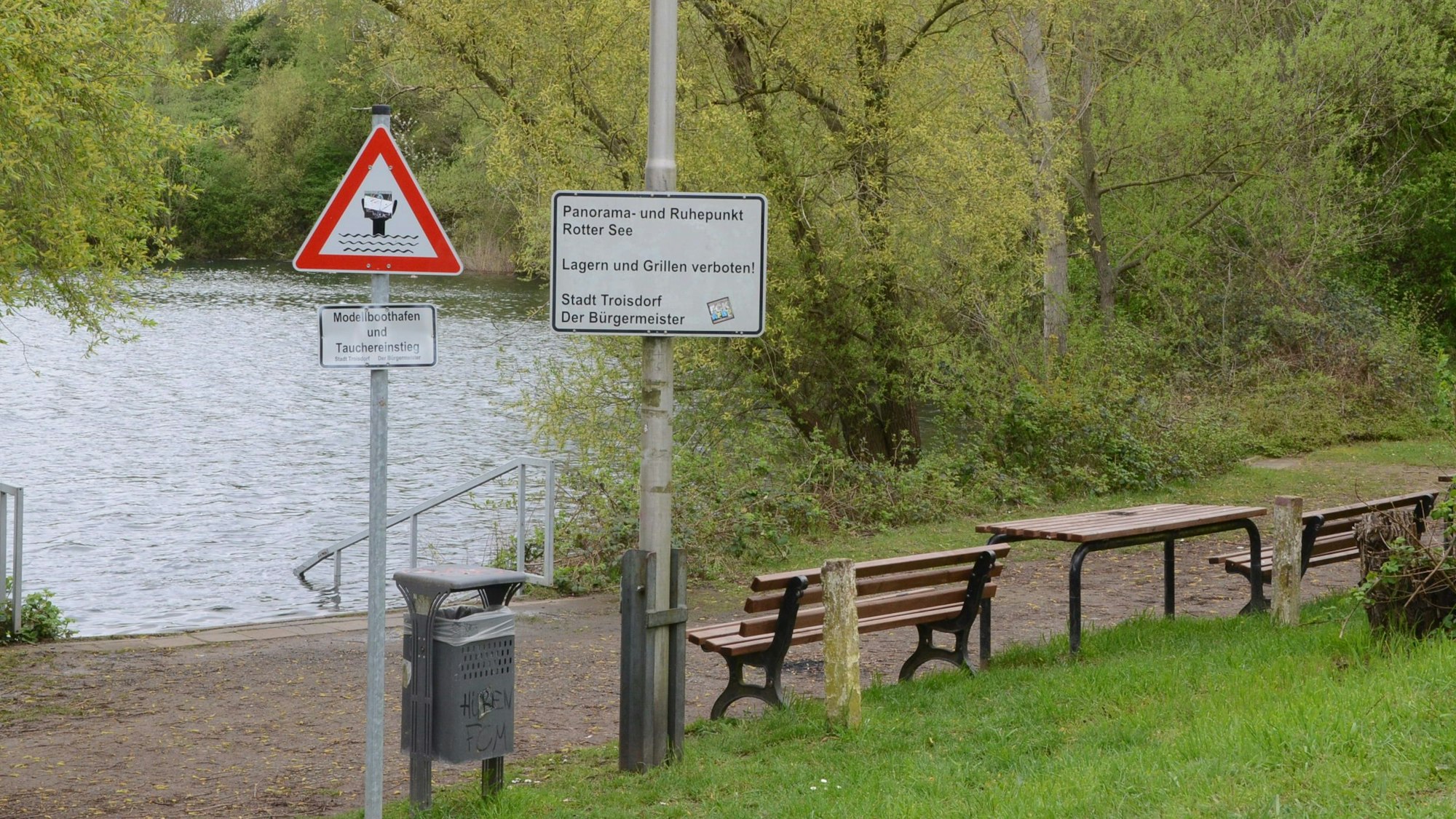 Fußweg an einem See; Bänke, ein Tisch und ein Papierkorb. Auf einem Schild steht „Panorama- und Ruhepunkt Rotter See“.