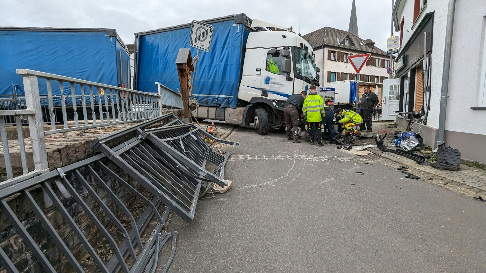 Ein Lastwagen ist in Hellenthal in ein Haus gefahren. Sachverständige und Polizisten schauen sich den Schaden an. Im Vordergrund liegen Brückengeländer-Elemente.