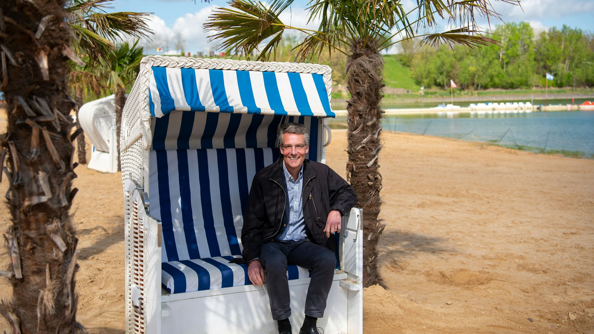 Geschäftsführer  Christoph Hartmann sitzt in einem Strandkorb unter Palmen am Badestrand des Zülpicher Sees.