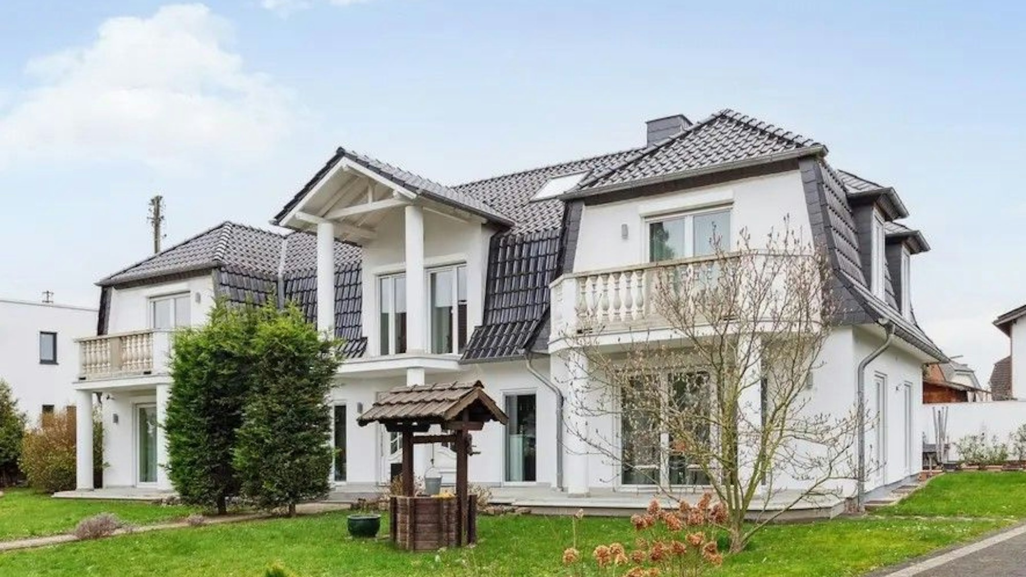 Ein weißes Einfamilienhaus mit dunkel gedecktem Dach, davor Rasenfläche und mehrere Pflanzen.