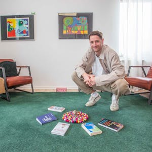 Therapeut Matthias Stinn in der Hocke auf einem grünen Teppich, vor sich hat er Bücher liegen.