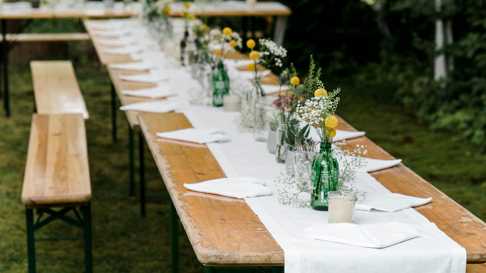 Zu sehen ist das Tischarrangement einer nachhaltigen Gartenhochzeit, mit Bierbänken und Zelten. Der Tisch ist geschmückt mit weißer, dezenter Stoffdeko und Wiesenblumen.