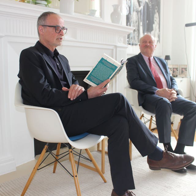 Zwei Männer sitzen auf Stühlen, einer liest aus einem Buch.