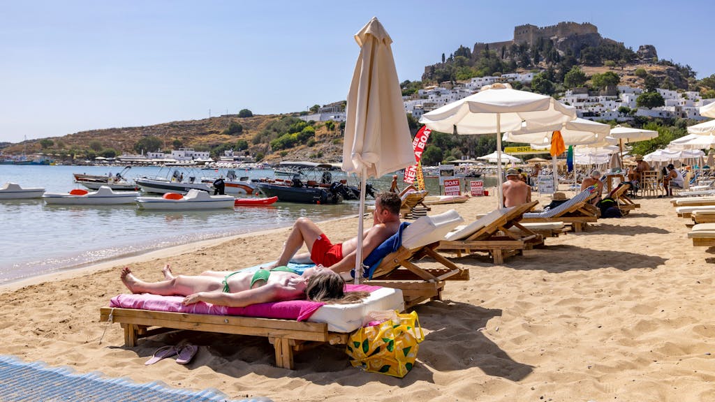 Am Strand in Lindos, etwa 10 Kilometer von den Bränden im Südosten entfernt, sind weiterhin Touristen am Strand, im Hintergrund die weltbekannte Akropolis von Lindos.
