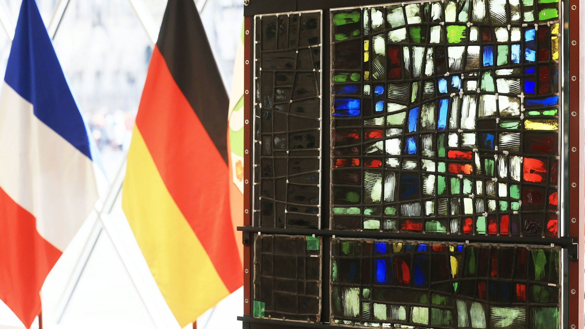 Der restaurierte (r.) und verrußte Teil (l.) eines Fenster aus der Kirche Notre-Dame hängen neben der deutschen und französischen Fahne im Kölner Domforum.Die Wiederherstellung der Glaskunst wurde als Symbol deutsch-französischer Zusammenarbeit verstanden.