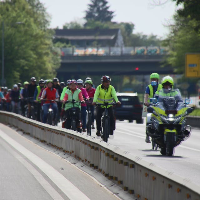 Ein dichter Pulk von Fahrradfahrern, begleitet von Polizei auf einem Motorrad.&nbsp;
