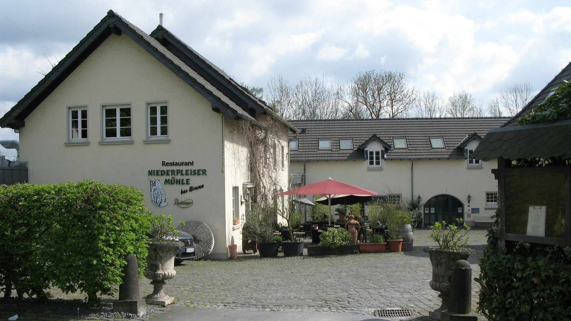 Das Foto zeigt den Zugang in den grob gepflasterten Hof der Niederpleiser Mühle mit hellgelb gestrichen Gebäuden. Vor dem Eingang stehen Pflanzkübel und ein roter Sonnenschirm.