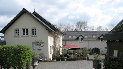 Das Foto zeigt den Zugang in den grob gepflasterten Hof der Niederpleiser Mühle mit hellgelb gestrichen Gebäuden. Vor dem Eingang stehen Pflanzkübel und ein roter Sonnenschirm.