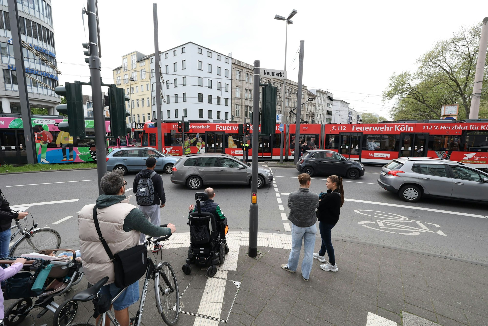 Fußgänger und Radfahrende warten an einer Ampel am Kölner Neumarkt darauf, dass sie die Straße überqueren können. Es sind Autos zu sehen und eine lange Straßenbahn.