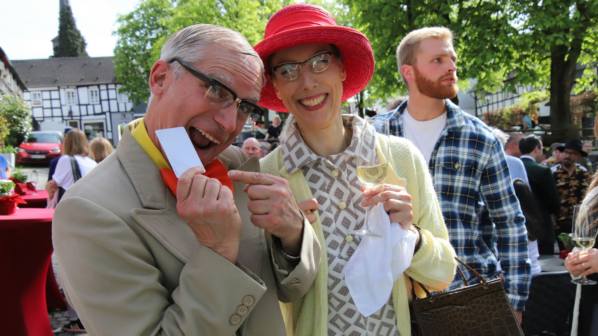 Ein Mann mit hellem Anzug und roter Fliege und eine Frau mit rotem Hut und Sektglas in der Hand posieren mit einer Zugangskarte für das Hotel.